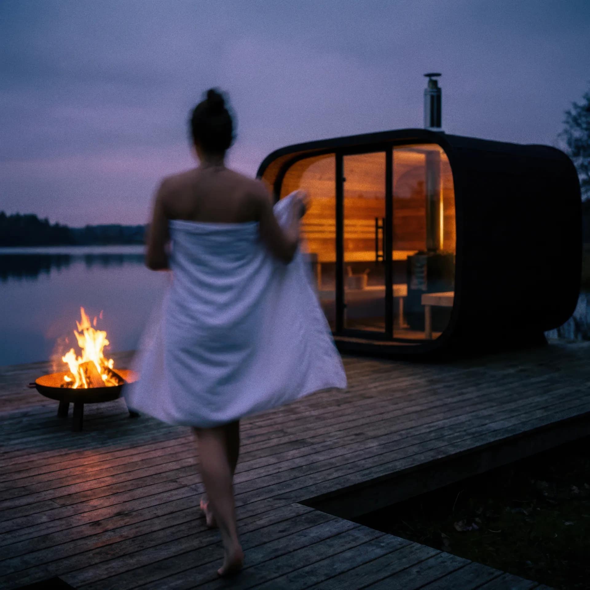 Sauna extérieur Round Cube au bord d’un lac au crépuscule, brasero allumé sur la terrasse